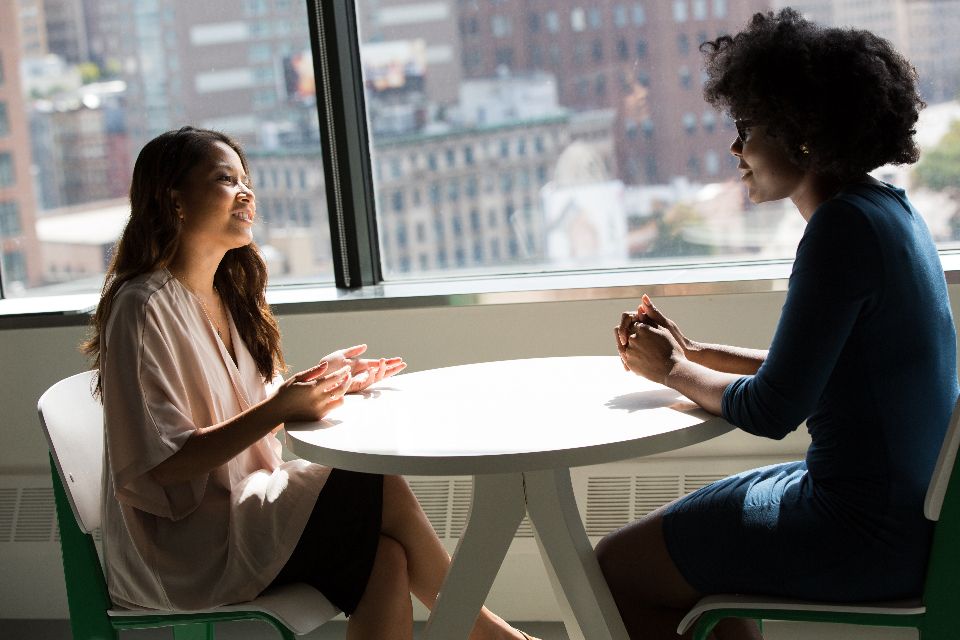 Two women talk across a small round table by a large window.