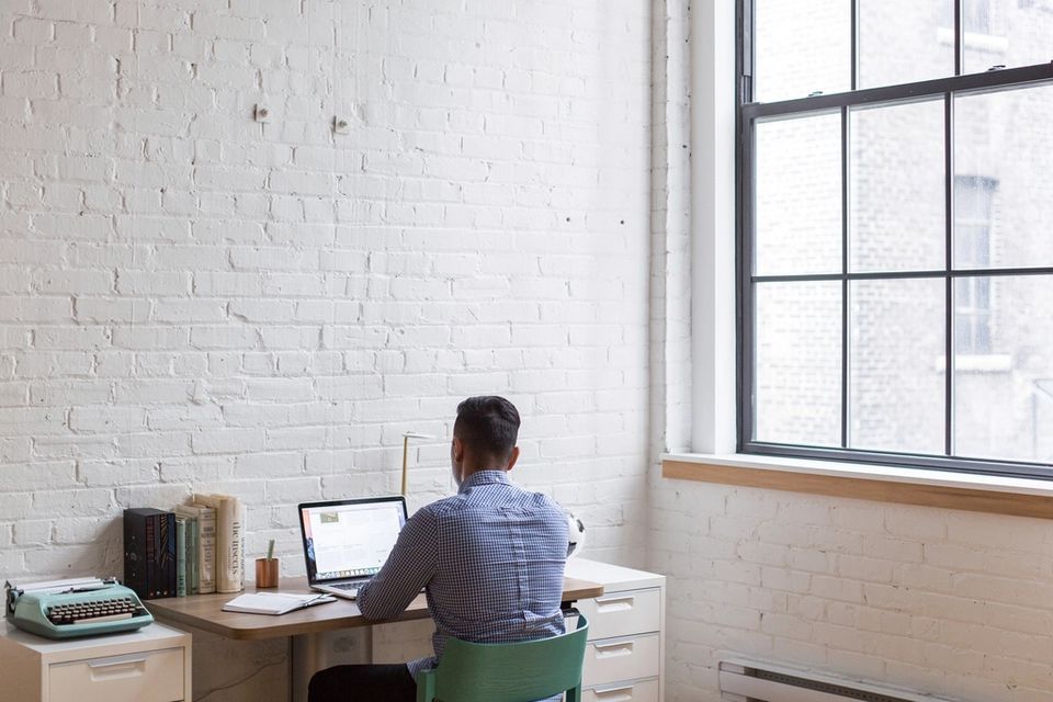 Un homme travaille sur un portable à un petit bureau, devant un mur de brique blanche et une grande fenêtre.